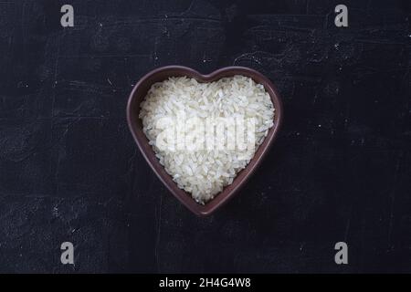 Une poignée isolée de riz blanc cru dans le bol en bois en forme de coeur sur fond de pierre noire Banque D'Images