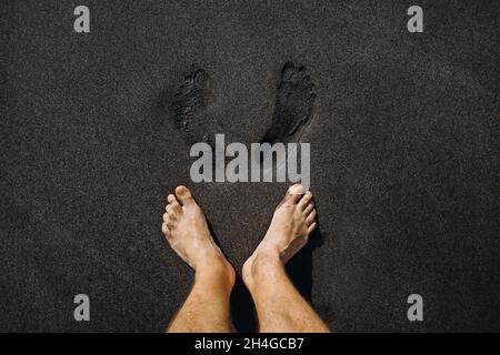 Gros plan des empreintes de pas mâles et des pieds marchant sur le sable noir volcanique de la plage. Banque D'Images