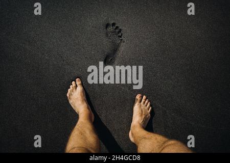 Gros plan des empreintes de pas mâles et des pieds marchant sur le sable noir volcanique de la plage. Banque D'Images