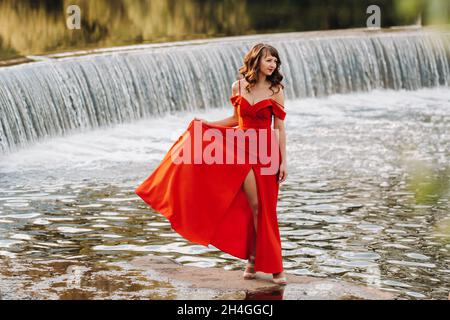 Fille dans une longue robe rouge près du lac dans le parc au coucher du soleil Banque D'Images