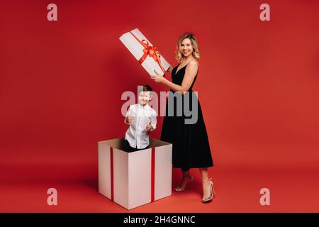 Maman ouvre un énorme cadeau de Noël dans lequel un garçon se tient et montre la classe sur un fond rouge. Banque D'Images