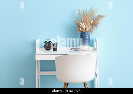 Table avec des pointes séchées dans un vase, réveil, tasse de café et lunettes près du mur bleu Banque D'Images