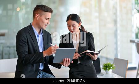 Un mentor de patron masculin serviable explique la campagne de stratégie marketing aux jeunes femmes asiatiques qui sont employées par le biais d'une tablette numérique au bureau. Banque D'Images