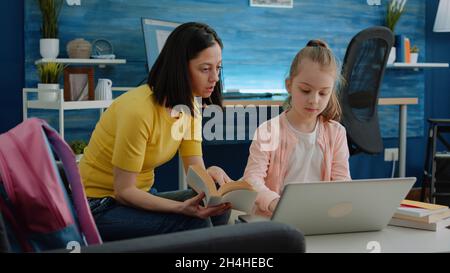 Mère et écolière lisant le livre et faisant des devoirs ensemble pour des leçons et des cours en ligne.Petit enfant utilisant un ordinateur portable, recevant l'aide de parents avec le travail scolaire et l'éducation à distance. Banque D'Images