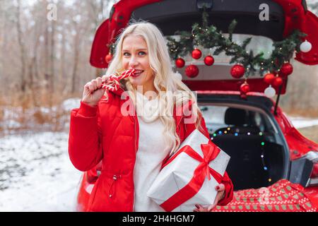 Femme blonde souriante en blouson rouge avec des cannes rouges et blanches en forme de coeur tenant une boîte cadeau avec ruban rouge près de la voiture décorée de noël en hiver Banque D'Images