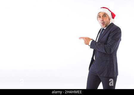 Homme d'affaires habillé en costume, cravate et chapeau de Noël du Père Noël, surpris, pointant avec ses mains, sur fond blanc.Fête de Noël conc Banque D'Images