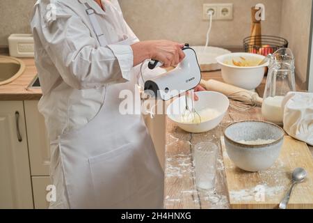 Une femme adulte vêtue de chef blanc bat des œufs avec un mélangeur Bosch pour faire une tarte dans la cuisine maison - Moscou, Russie, 2 septembre 2021 Banque D'Images