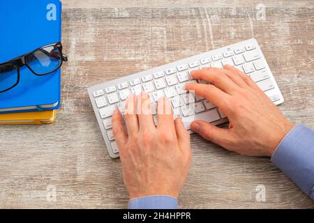 Main de l'homme tapant sur le clavier blanc sur le bureau en bois. Rapport d'entreprise, vue de dessus Banque D'Images