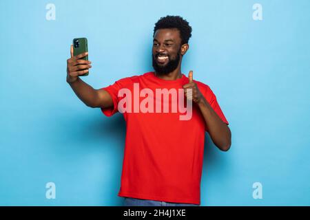 L'homme afro-américain fait des selfies gesturant des thumbs en posant sur fond bleu.Photo en studio Banque D'Images