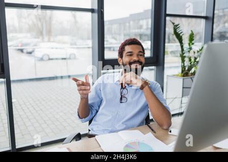 Homme d'affaires américain africain positif tenant des lunettes et pointant avec le doigt près de l'ordinateur dans le bureau Banque D'Images