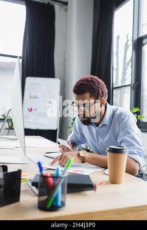 Homme d'affaires afro-américain tenant un smartphone et pointant vers un document près d'un ordinateur au bureau Banque D'Images