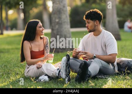 Couple assis sur l'herbe d'un parc buvant une boisson non alcoolisée portant des patins en ligne Banque D'Images