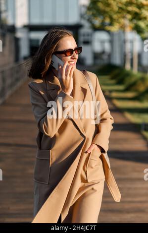 Élégante femme heureuse en manteau d'automne et lunettes de soleil marchant à l'extérieur et parlant sur téléphone portable, gaie femme parlant et bavardant par téléphone mobile dans la rue, marchant dans le parc Banque D'Images