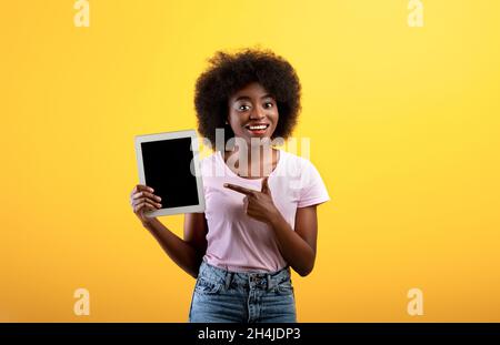Bonne dame afro-américaine pointant vers une tablette numérique avec écran vierge, fond jaune, maquette Banque D'Images