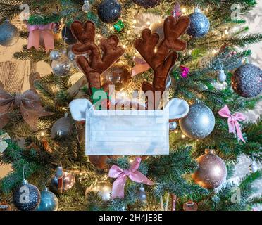 Joyeux Noël dans la nouvelle réalité Covid-19.Vue de face d'un arbre de Noël avec masque médical blanc protecteur et oreilles de renne décorées Banque D'Images