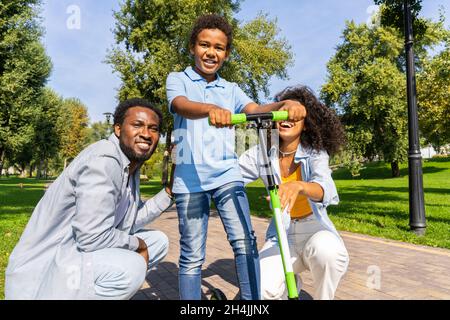 Belle famille afro-américaine heureuse de se lier au parc - famille noire ayant l'amusement à l'extérieur, parents enseignant à son fils à monter sur le scooter Banque D'Images