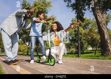 Belle famille afro-américaine heureuse de se lier au parc - famille noire ayant l'amusement à l'extérieur, parents enseignant à son fils à monter sur le scooter Banque D'Images