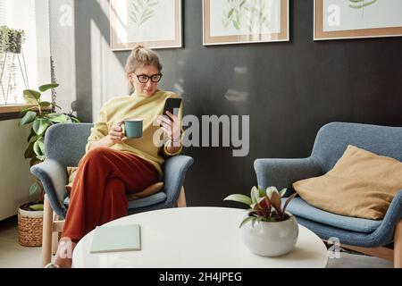 Jeune femme en lunettes assise sur un fauteuil avec un téléphone portable et buvant du café dans la chambre Banque D'Images
