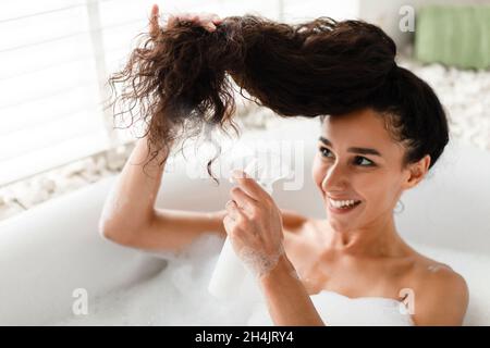 Traitement de réparation des extrémités fractionnées.Jeune femme souriante appliquant un spray à l'huile essentielle sur ses cheveux tout en étant allongé dans un bain moussant à la maison.Belle femme millénaire m Banque D'Images
