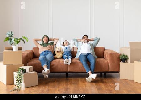 Une jeune famille du Moyen-Orient satisfaite s'est penchée sur le canapé, se reposant après avoir été déplacé, assis près des boîtes en carton et des plantes dans un nouvel appartement, copie libre de la flèche Banque D'Images