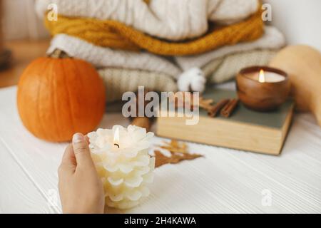 Main tenant la bougie sur fond de citrouille, sweaters confortables, feuilles d'automne et livre vintage sur fond de bois blanc dans la chambre.Bonjour automne, Banque D'Images