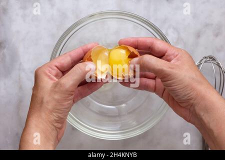 Vue de dessus de la femme craquant à la main oeuf brun cru dans un bol en verre sur la surface en marbre Banque D'Images