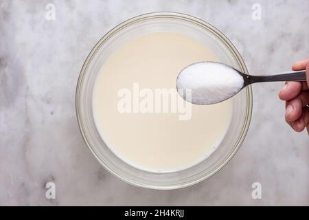 Vue de dessus d'une femme tenant à la main une cuillère de sucre et mettant dans la pâte dans un bol en verre sur la surface en marbre Banque D'Images