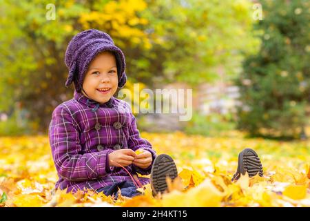 Petite fille caucasienne de cinq ans assise sur terre avec des feuilles jaunes dans le parc d'automne Banque D'Images