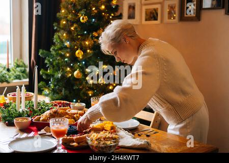 Bonne jeune femme préparant Noël table de dîner à la maison pour la fête de famille sur un fond flou de décoration arbre de Noël et des lumières de célébration. Banque D'Images