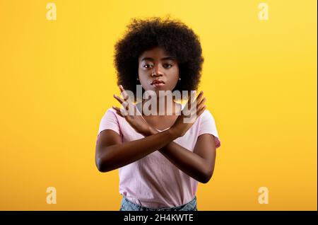 Concept de refus.Mécontent femme noire montrant un geste d'arrêt avec des mains croisées, refusant quelque chose de non désiré en se tenant sur fond jaune studio Banque D'Images