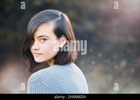 Contenu bonne adolescente posant regardant l'appareil photo au-dessus de son épaule.Reflets de l'objectif avec un arrière-plan automnal flou. Banque D'Images