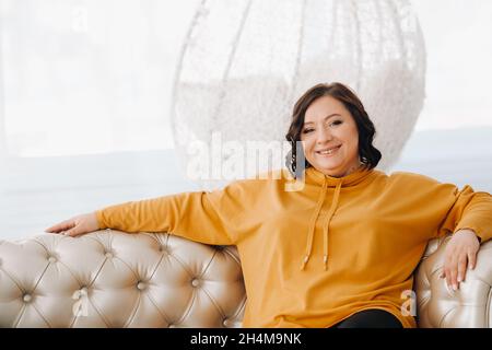 une femme à capuche orange est assise sur un canapé blanc. Banque D'Images