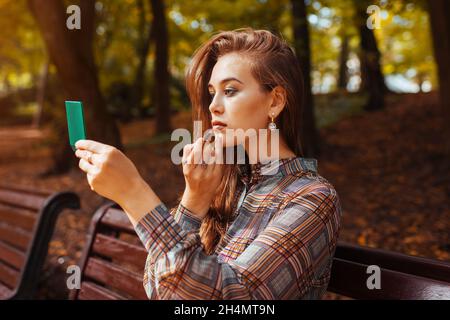 Portrait d'une jeune femme appliquant un rouge à lèvres à l'aide d'un miroir de la main dans un parc d'automne assis sur un banc.Fille vérifiant le maquillage à l'extérieur Banque D'Images