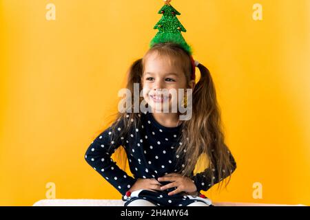 Portrait de sourire positif surpris bonne petite écofille fille Noël arbre décoration sur la tête polka robe à pois montrer pouces vers le haut fond orange Banque D'Images