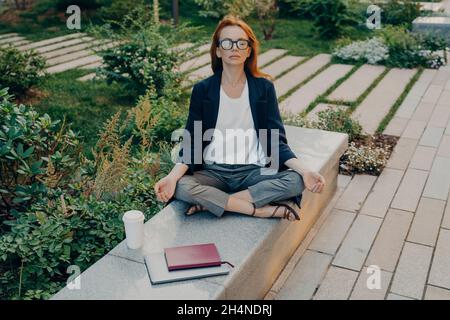 REDHEAD la jeune femme européenne est assise en position lotus porte des vêtements formels boissons à emporter café Banque D'Images