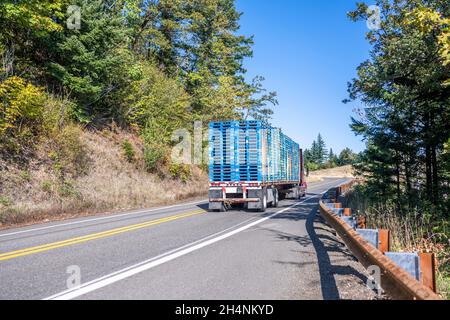 Puissant camion semi-lourd rouge classique transportant des palettes en bois empilées fixées sur un semi-remorque à lit plat grimpant sur la route sinueuse de montagne wi Banque D'Images