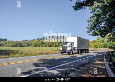 Semi-camion classique à capot blanc avec cabine de jour transportant des marchandises commerciales dans une remorque semi-remorque à fourgon sec fonctionnant pour livraison sur le bobinage étroit Banque D'Images