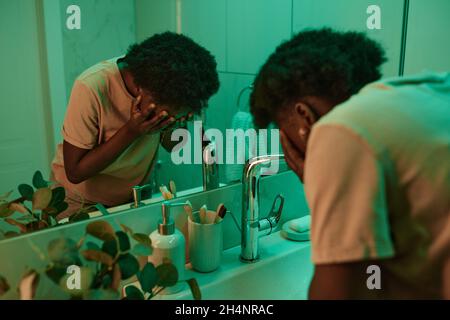 Une jeune femme africaine lavant son visage avec de l'eau avant de dormir debout dans la salle de bains en face du miroir Banque D'Images