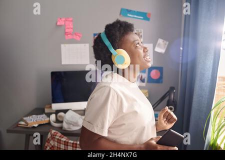 Jeune femme africaine écoutant de la musique dans des écouteurs sans fil et dansant dans la chambre Banque D'Images