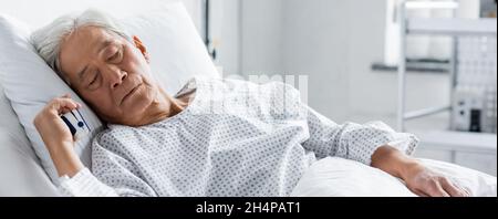 Patient asiatique âgé avec oxymètre de pouls dormant au lit en clinique, bannière Banque D'Images