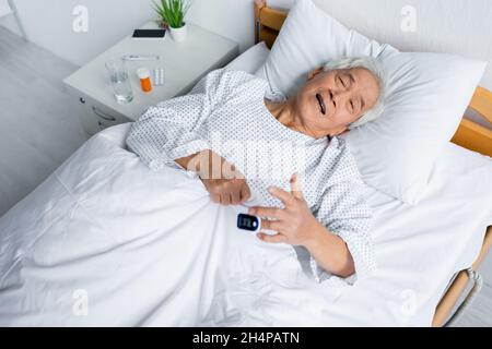Vue en grand angle d'un patient asiatique malade avec un oxymètre de pouls allongé sur le lit près des pilules en clinique Banque D'Images