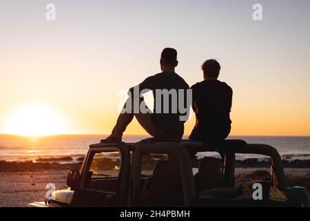 Vue arrière d'un couple de hommes gais caucasiens assis sur le toit de la voiture au coucher du soleil au bord de la mer Banque D'Images