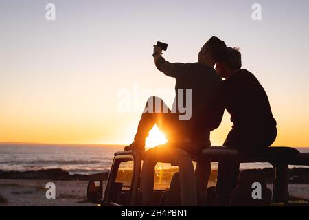 Vue arrière d'un couple de sexe masculin gay caucasien assis sur le toit d'une voiture prenant des selfies au coucher du soleil au bord de la mer Banque D'Images