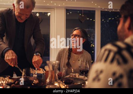 Homme senior servant de la nourriture à sa famille assis à table le soir de Noël.Repas de Noël en famille. Banque D'Images