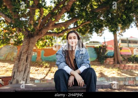 Une adolescente confiante qui regarde l'appareil photo à l'extérieur.Une fille brune insouciante portant une veste en denim dans la ville.Jeune femme assise seule Banque D'Images