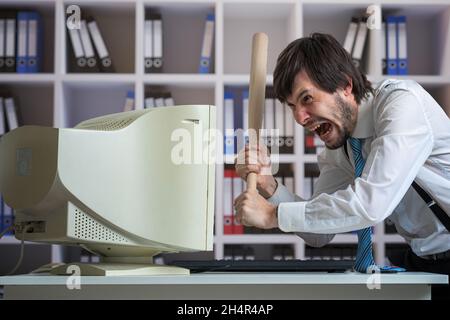 Un homme en colère détruit un vieux ordinateur avec une batte de baseball au bureau. Banque D'Images