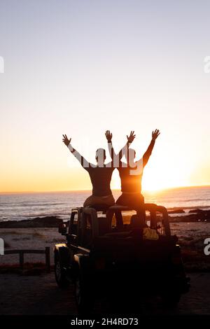 Vue arrière d'un couple de sexe masculin gay caucasien assis sur le toit d'une voiture levant les bras au coucher du soleil au bord de la mer Banque D'Images