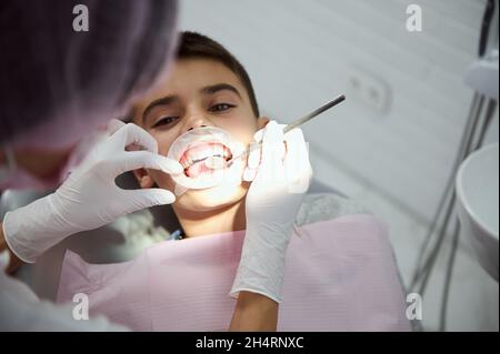 Gros plan d'un petit garçon portant un écarteur de joue dentaire lors d'un examen médical régulier.Vue en hauteur du dentiste à l'aide d'un miroir dentaire en acier inoxydable Banque D'Images