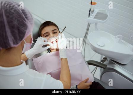 Vue du dessus du dentiste pédiatre traitant les dents de l'enfant.Gros plan d'un garçon âgé d'âge élémentaire avec une bouche ouverte et un enrouleur de joue à la dentition dentaire Banque D'Images