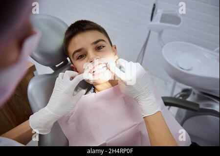 Gros plan d'un joli petit garçon d'école qui regarde un appareil photo avec un sourire éclatant et des dents blanches saines tandis que le dentiste pédiatrique met un chien Banque D'Images